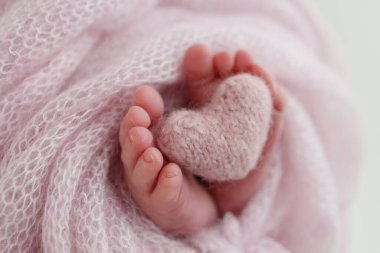 Knitted pink heart in the legs of a baby. Soft feet of a new born in a pink wool blanket. Close-up of toes, heels and feet of a newborn. Macro photography the tiny foot of a newborn baby.