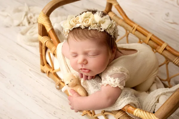 白いスーツを着た可愛い新生児と 頭に花をつけたヘッドバンドが甘い眠りにつく ウィッカーは白いリネンでベッドをガラガラ ライトウッドを背景にしたプロ仕様のマクロ写真 — ストック写真