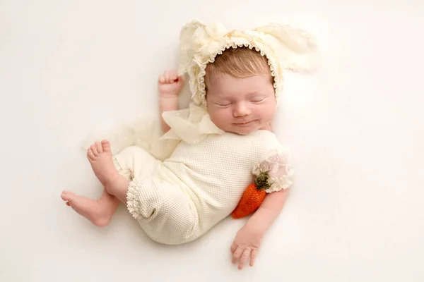 白いスーツのかわいい新生児とウサギの耳の帽子は甘く眠ります 赤いフェルトのニンジンの手に 白い背景にウサギのプロの写真のクローズアップのイメージ — ストック写真