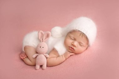 Beyaz tulumla uyuyan, pembe arka planda beyaz şapkalı yeni doğmuş bir kızın üst görüntüsü. Pembe oyuncak tavşanla. Küçük bir bebeğin güzel portresi. 7 gün, 1 haftalık.