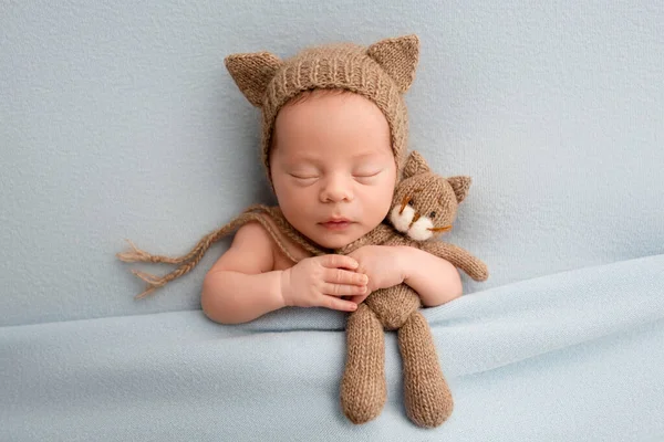 A cute newborn boy in the first days of life sleeps in a brown hat with cat ears. On a blue background with a blue blanket. With a toy brown kitten. High quality photo
