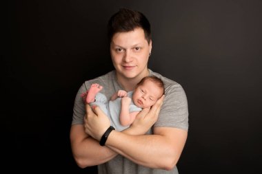 Yeni doğmuş şirin bir erkek bebek, hayatın ilk günlerinde mavi tulumla uyur. Babamızın kollarında. Sıcak ebeveyn sarılması. Siyah bir arka planda. Profesyonel portre fotoğrafı.. 