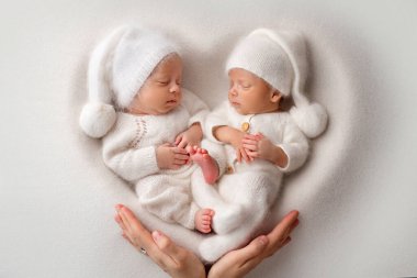 Tiny newborn twin boys in white bodysuits on a white background. Newborn twins sleep next to their brother on the background of the heart. Parents mother holds children with their hands with palms. 