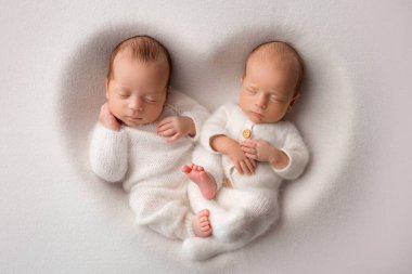 Tiny newborn twin boys in white bodysuits on a white background. Newborn twins sleep next to their brother on the background of the heart. Two newborn twin boys hugging each other. High quality photo