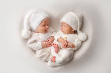 Tiny newborn twin boys in white bodysuits on a white background in white caps. Newborn twins sleep next to their brother on the background of the heart. Two newborn twin boys hugging each other. 