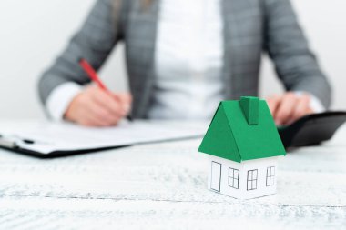 Businesswoman sitting and writing in notebook. Paper house on desk.