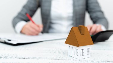 Businesswoman sitting and writing in notebook. Paper house on desk.
