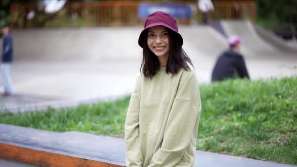 帽子の中の緑色のセーターを着たブルネットの女の子の肖像画は スケートパークの背景に座ってカメラのポーズをとっています レジャーを歩く — ストック動画