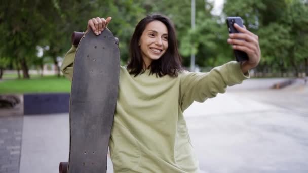 緑のセーターを着たブルネットの肖像画 彼女はスケートボードを手に公園の背景に座って自撮りをします 自撮り レジャーを歩く — ストック動画