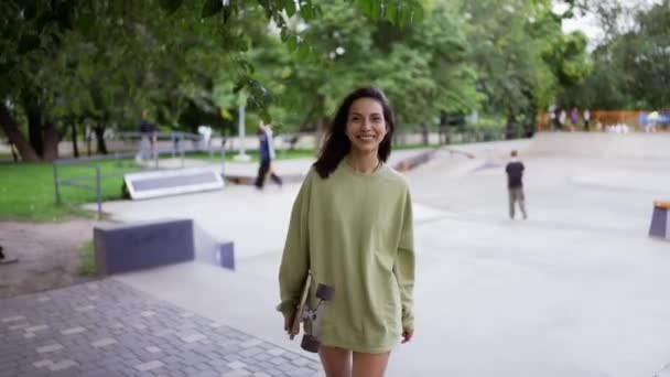 緑のセーターを着た幸せなブルネットの女の子が公園でスケートボードと笑顔で歩いています 公園を歩きなさい 肖像画 レジャー — ストック動画
