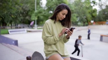 Yeşil kazaklı esmer bir kız kaykay parkının arka planında oturuyor ve telefonda mesajlaşıyor. Parkta yürüyüş, hobi, eğlence..