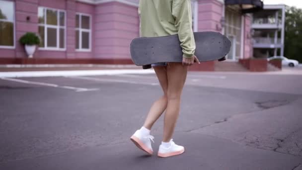 女の子はスケートボードを手に公園を歩く 毎日の趣味 公園を歩き スケートをスケートボーダーパークに運ぶ — ストック動画