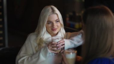 Beautiful long-haired blonde girl is chatting with her friend over a cup of cappuccino at the cafe.