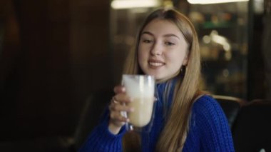 Smiling woman holds big transparent cup of cappuccino in hands then drinks.