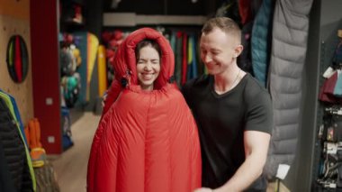 A smiling couple purchase in sportswear store, trying sleeping bag and make selfie.