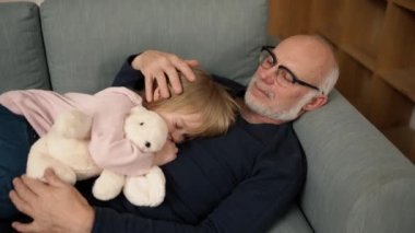 Little girl with toy sleeping on his grandfathers chest in the living room.