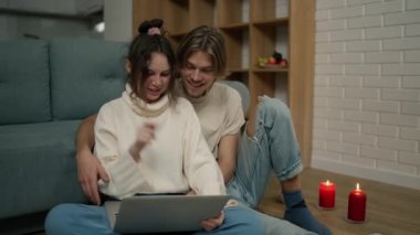 Young adult couple using laptop computer at home sitting on floor around candles.