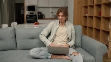 Relaxed man sitting on sofa working from home on laptop.
