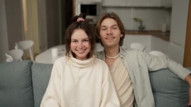 Smiling millennial couple home owners sitting on cozy sofa, showing keys to camera, feeling excited.