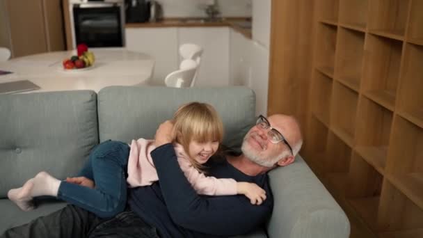 孫娘は彼女の祖父の上にソファの上にジャンプし 彼を抱擁 — ストック動画