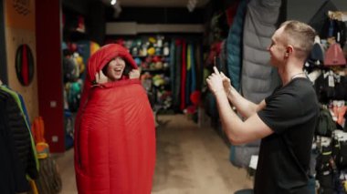 A smiling couple purchase in sportswear store taking pictures inside the sleeping bag.