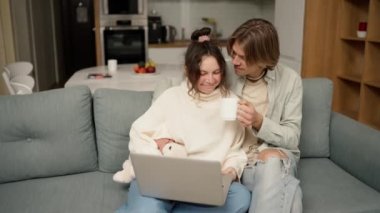 Young adult couple talking using laptop computer at home sitting on couch, watching video.