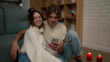 Romantic couple sitting on the floor together, looking on smartphone screen.