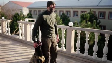 A man stands on terrace with service dog in military protective collar.