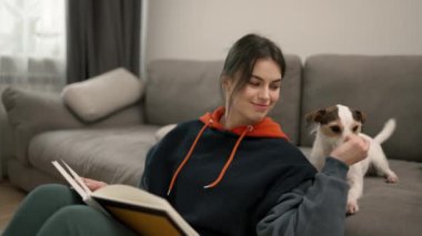 A beautiful woman reading book and caress her pet dog on the couch at home.