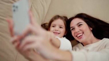 Mother and daughter taking selfie at home while laying on bed.