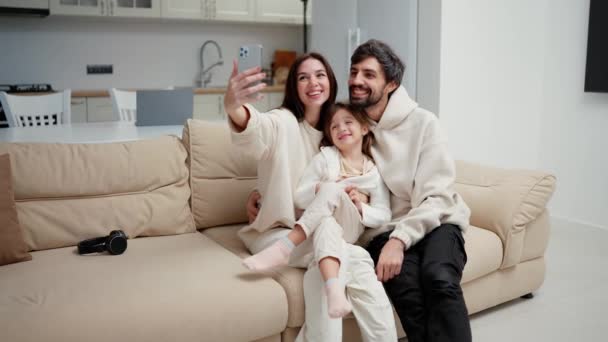 幸せな笑顔の家族はリビングルームでソファの上にスマートフォンで自撮りやビデオ通話をしています — ストック動画