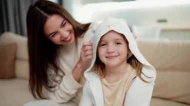 Loving mother embracing kid daughter and putting hood on the head while sitting on sofa.