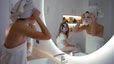 Mother applying tissue panda mask on her face relaxing in the bedroom with her daughter next to her.