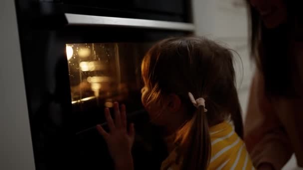 小さな女の子はガラスを通してどのようにクッキーがオーブンで焼かれるか見ています — ストック動画