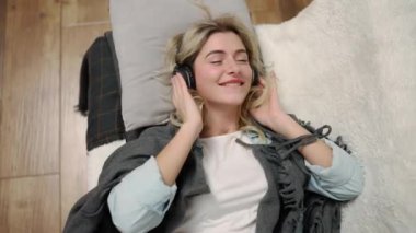 A smiling woman falls on a bed in headphones and listens to music.