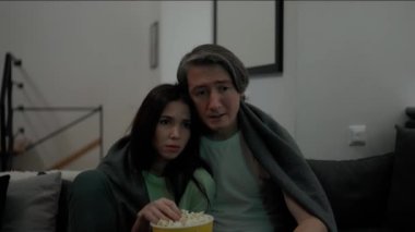 Couple sitting on sofa eating popcorn and watch thriller or drama on TV together.