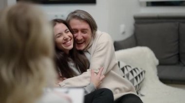 Over shoulder - couple visiting psychotherapist office, a man hugs his wife, both smiling.