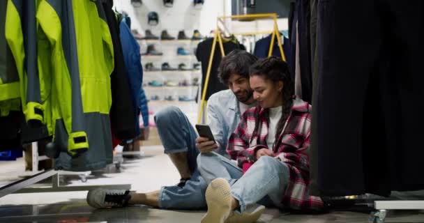 体育百货商店里的情侣们坐在地板上 看着手机 — 图库视频影像