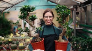 Çiçekçide ya da serada çiçek sıraları arasında yürürken elinde bitkilerle gezen önlüklü genç bayan çiçekçi..