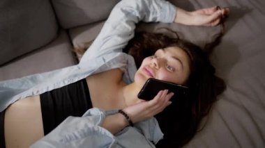 Uzun saçlı kadın kanepeye uzanıp cep telefonu kullanıyor, dinleniyor..