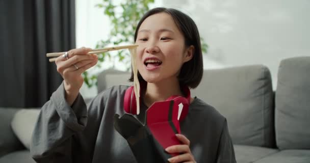 若いですアジアの女性でヘッドフォン食べる麺とともに木の棒で家 — ストック動画
