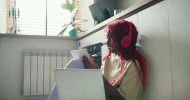Kırmızı kıvırcık saçlı Afro-Amerikalı kız mutfakta dizüstü bilgisayarla ders çalışıyor, notlar alıyor..