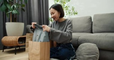 Şirin Asyalı kadın yeni kıyafetlerle kese kağıdını açıyor. Oturma odasında yerde oturuyor..