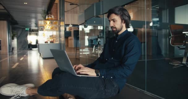成功的办公室职员舒适地在笔记本电脑上工作 办公室职员坐在玻璃墙旁边的地板上 用笔记本电脑工作 — 图库视频影像