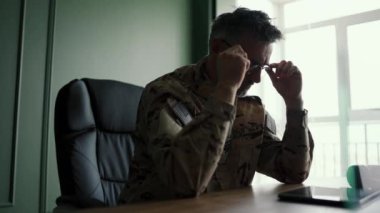 Kamuflajlı bir asker masaya oturur ve şeffaf gözlüklerini çıkarır. Bir askerin işyeri..