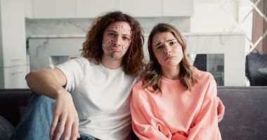 Çift evde bir kanepede oturuyor, stresli ve rahatsız, olumsuz ruh hali yüzünde yapışkan bandajlarla.