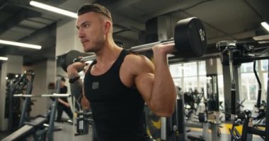 Siyah gömlekli genç bir atlet bacak kaslarını geliştirmek için halterle çömeliyor. Spor salonunda bacak günü, bacak kaslarını güçlendirmek için egzersiz..