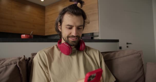 年轻的布鲁内特男子在红色长椅上用无线耳机玩手机游戏 — 图库视频影像