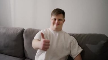 Kanepede oturan genç bir adam, mutlu bir şekilde gülümsüyor. Tamam işareti yapıyor, parmaklarıyla başparmağını kaldırıyor, mükemmel bir işaret..