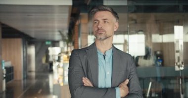 Gri saçlı, gri takım elbiseli, kendinden emin orta yaşlı bir adam ofisin arka planında poz veriyor. Ellerini göğsüne koy..